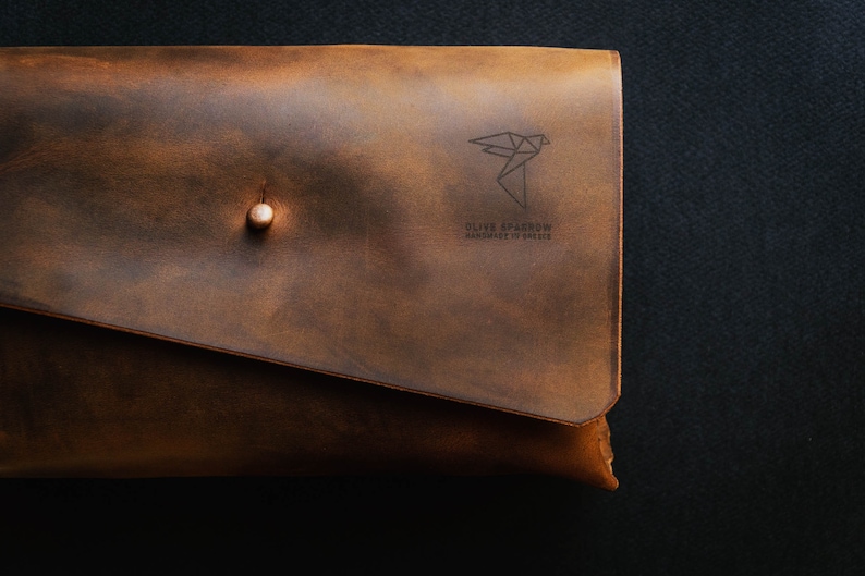 May include: A brown leather clutch with a brass button closure. The clutch has a geometric bird design and the text "Olive Sparrow Handmade in Greece" embossed on the front.