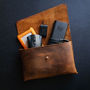 May include: A brown leather pouch with a snap closure, containing a black mouse, a black Dell power adapter, a black USB drive, and an orange external hard drive.