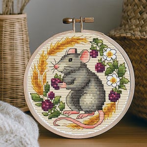 Könnte beinhalten: Kreuzstich-Stickerei einer grauen Ratte, die Beeren hält, umgeben von Weizen und Blumen, in einem Holzreifen. Das Kunstwerk wird auf einer Holzoberfläche mit einer gewebten Vase und einem Korb im Hintergrund ausgestellt.