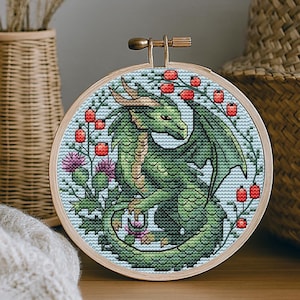 Può includere: Un telaio da ricamo a punto croce con un drago verde con ali dettagliate, circondato da bacche rosse e cardi viola. Il drago è su uno sfondo azzurro, incorniciato in un cerchio di legno.