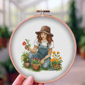 Op de afbeelding: Een kruissteek borduurring met een vrouw in een overall en hoed die bloemen in potten verzorgt. Het ontwerp bevat oranje en rode bloemen, een troffel en een houten ring. De achtergrond is een kas.