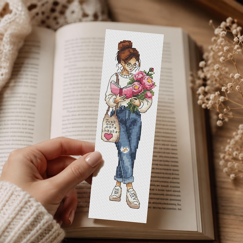 Op de afbeelding: Een kruissteek bladwijzer met een illustratie van een persoon die een boek en een boeket roze bloemen vasthoudt. De persoon draagt een bril, jeans en sneakers en draagt een draagtas met de tekst "Just one more chapter".