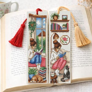May include: Two rectangular cross-stitch bookmarks featuring illustrations of people reading. One bookmark has a red tassel and shows a person by a window with a dog. The other has a beige tassel and shows a person with a cat, bookshelves, and a plant.