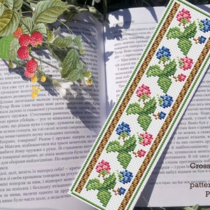 Op de afbeelding: Geborduurd bladwijzer met een bloemmotief van roze en blauwe bessen, groene bladeren en een bruine rand. De bladwijzer ligt op een open boek met tekst en een takje frambozen.