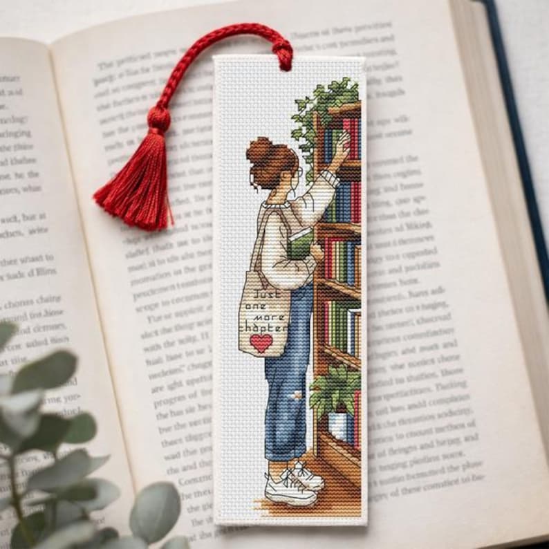 Op de afbeelding: Bladwijzer met een rode kwast, met een kruissteekontwerp van een persoon die naar een boek in een plank reikt. De persoon draagt een spijkerbroek en sneakers en draagt een draagtas met de tekst "Just one more chapter."