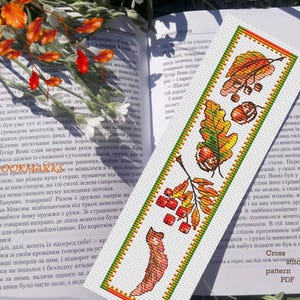 May include: A handmade bookmark with a cross-stitch design featuring autumn leaves, acorns, and berries in shades of orange, green, and brown. The bookmark is on a white background with a green border.