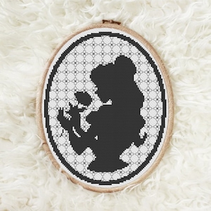 May include: Black and white cross-stitch embroidery of a silhouette of a woman holding a flower in a circular wooden frame.