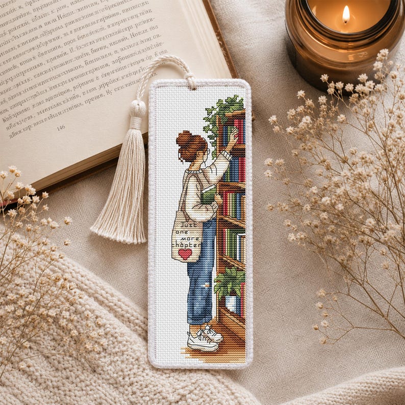 Op de afbeelding: Geborduurd boekenlegger met een vrouw die naar een boek in een plank reikt. De boekenlegger heeft een kwast en de tekst "Just one more chapter" op een draagtas. Het kleurenpalet omvat wit, blauw, groen en bruin.