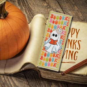 May include: A white ghost with a flower on its head is holding a book. The ghost is on a bookmark with a colorful pattern. The bookmark is on an old book with the text "Happy Thanks Giving".