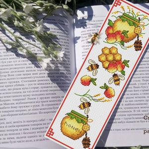 Op de afbeelding: Een kruissteek-bladwijzerpatroon met een honingpot, honingraat, appels en bijen. Het patroon is op witte stof met rode en gele accenten. De tekst "Honey" is geborduurd op de honingpot. De tekst "Cross stitch pattern PDF" staat onderaan de bladwijzer.