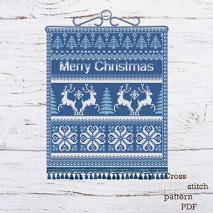 May include: A blue and white cross-stitch pattern with the words "Merry Christmas" in the center. The design includes reindeer, snowflakes, and Christmas trees. The bottom edge has a fringe of tassels. The pattern is suspended from a decorative metal hanger.