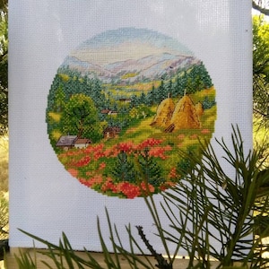 Op de afbeelding: Een cirkelvormig kruissteek borduurwerk op witte Aida-stof met een landelijk landschap met kleine huisjes, groene bomen, gouden hooibergen en een veld met rode bloemen onder een zachte blauwe lucht met verre heuvels op de achtergrond.