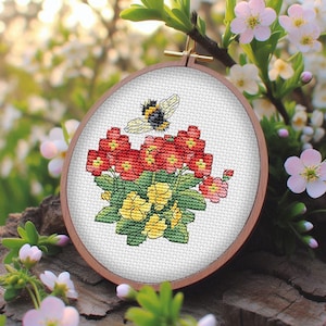 May include: An oval cross-stitch embroidery in a wooden hoop depicts a floral design with red and yellow flowers, green leaves, and a bumblebee. The background is blurred, showing flowers and greenery.