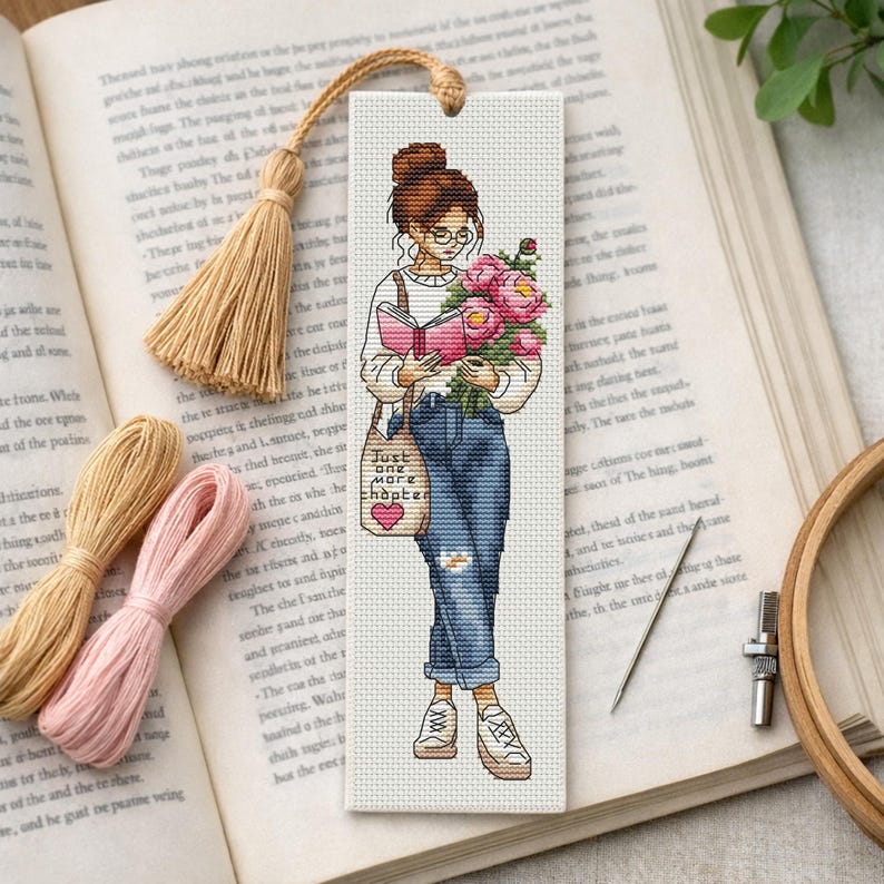 Op de afbeelding: Kruissteek bladwijzer met een vrouw die een boek leest en bloemen vasthoudt. De bladwijzer heeft een kwast en de tekst "Just one more chapter". De vrouw draagt een bril, jeans en sneakers.