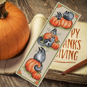 May include: A bookmark with a Thanksgiving theme featuring three black cats sitting on top of pumpkins. The bookmark is made of fabric and has a beige background with green and orange accents. The text "Happy Thanksgiving" is printed on the bookmark.