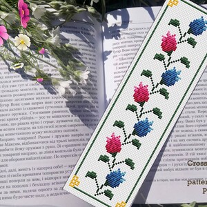 Könnte beinhalten: Weißes Kreuzstich-Lesezeichen mit einem floralen Muster in Rosa und Blau. Das Lesezeichen hat einen grünen Rand und liegt auf einem offenen Buch. Der Text "Cross stitch pattern PDF" ist sichtbar. Blumen im Hintergrund.