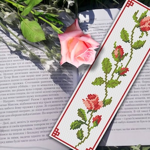 Op de afbeelding: Een witte bladwijzer met een rode rand heeft een kruissteekontwerp met drie rode rozen met groene bladeren en stelen. De bladwijzer ligt op een boek met een roze roos erop.