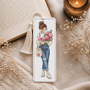 Op de afbeelding: Geborduurd boekenlegger met een vrouw die een boek en bloemen vasthoudt. Ze draagt een bril, jeans en sneakers. Een draagtas leest "Just one more chapter". De boekenlegger heeft een kwast en een zachte, neutrale achtergrond.