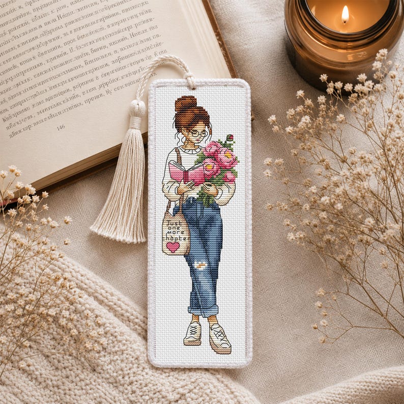 Op de afbeelding: Geborduurd boekenlegger met een vrouw die een boek en bloemen vasthoudt. Ze draagt een bril, jeans en sneakers. Een draagtas leest "Just one more chapter". De boekenlegger heeft een kwast en een zachte, neutrale achtergrond.