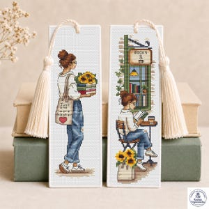 May include: Two rectangular white fabric bookmarks featuring cross-stitch designs. The left bookmark shows a person holding a stack of books with sunflowers and a tote bag reading "Just one more chapter". The right shows a person reading at a cafe.