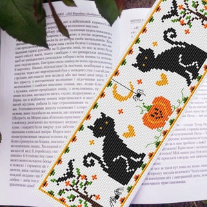 May include: A black and white cross-stitch bookmark featuring two black cats, a pumpkin, a spider, and a crescent moon. The bookmark is on a book with text in a foreign language.