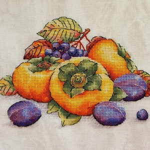 Puede incluir: Una bordado de punto de cruz que representa una naturaleza muerta con caquis y ciruelas. Los caquis son de color naranja y amarillo, y las ciruelas son de color morado. La fruta est&aacute; rodeada de hojas verdes.