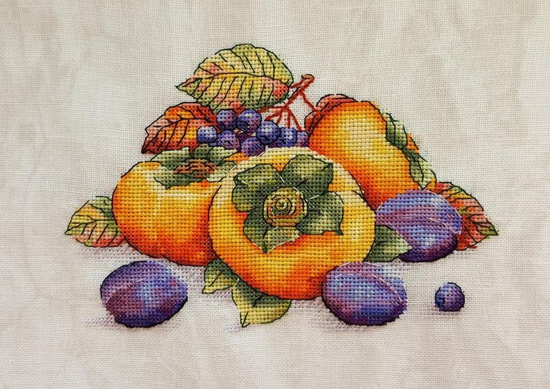 Puede incluir: Una bordado de punto de cruz que representa una naturaleza muerta con caquis y ciruelas. Los caquis son de color naranja y amarillo, y las ciruelas son de color morado. La fruta est&aacute; rodeada de hojas verdes.