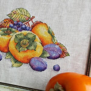 Puede incluir: Bordado de punto de cruz de un bodeg&oacute;n con caquis, ciruelas y uvas. La fruta est&aacute; dispuesta en un racimo con hojas verdes. El bordado est&aacute; enmarcado en un marco dorado.