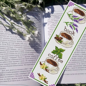 May include: A white cross-stitch bookmark with a coffee cup, a book, and a coffee cup design. The bookmark has the words "coffee" and "book" written on it.