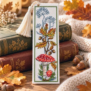 May include: A rectangular cross-stitch bookmark on white Aida cloth featuring a red and white spotted mushroom, autumn leaves, and acorns. The design is bordered by a thin green line and includes a cream-coloured tassel attached at the top.