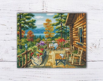 Puede incluir: Una cabaña acogedora con un porche de madera con vistas a un lago. El porche está decorado con flores coloridas y una mesa con sillas. Hay dos mecedoras en el porche y una mesa pequeña con un libro y una taza de café.