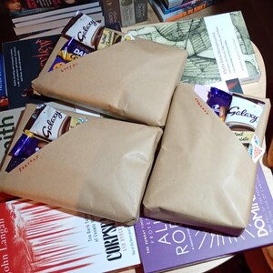 Op de afbeelding: Drie in bruin papier verpakte pakketten, elk met een chocoladereep en andere lekkernijen, liggen op een tafel omringd door boeken. De pakketten zijn gelabeld met de woorden "fantasy", "romance" en "horror".