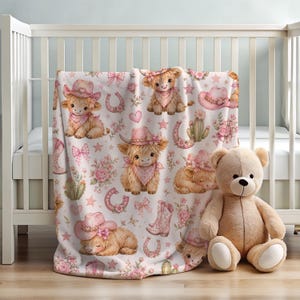 May include: A white baby blanket featuring a repeating pattern of light brown highland cows wearing pink cowboy hats and bandanas, interspersed with pink horseshoes, cacti, flowers, bows, and stars, draped over a white wooden crib.