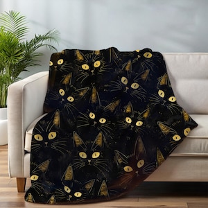 May include: A soft throw blanket featuring a repeating pattern of black cats with bright yellow eyes. The cats are depicted with fine gold-toned line details on their ears and whiskers, set against a dark, solid black background.