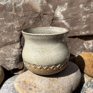 Puede incluir: Una maceta de cerámica hecha a mano con un esmalte gris claro moteado. La maceta tiene una forma redondeada con un borde ligeramente ensanchado y un diseño geométrico decorativo alrededor de la base. La maceta está sobre una piedra.