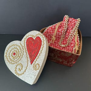 May include: Heart-shaped box with a red heart design on the lid. The lid is white with gold accents. Inside the box is a red fabric bag with a beaded drawstring. The box is open, revealing the contents.