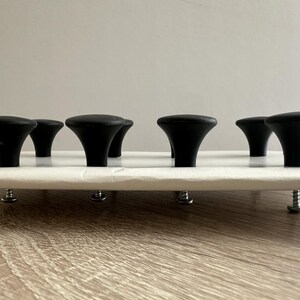 May include: A white, rectangular object with six black, mushroom-shaped knobs. The knobs are evenly spaced across the top surface. The object is elevated by silver screws, suggesting a furniture component or decorative piece.