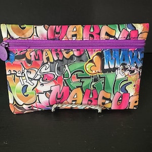 May include: A rectangular makeup bag with a vibrant graffiti design in multiple colors, including pink, green, and orange. The bag has a purple zipper and the word "Makeup" is visible in the design.