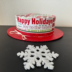 Op de afbeelding: Een rode en witte kerst hoed met de tekst "Happy Holidays! Joy to the World!" en een witte sneeuwvlok op een zwart oppervlak.