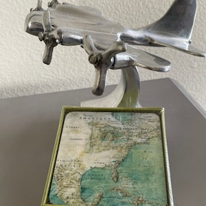 May include: A set of four coasters featuring a vintage map of North America with the text "AMERIQUE" and "GOLFE DE MEXIQUE". The coasters are packaged in a green box with a silver airplane figurine on top.