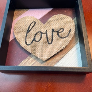 May include: A framed burlap heart with the word "love" written in black lettering. The heart is on a background of wood planks painted in shades of pink, red, brown, and white.