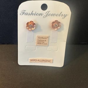 May include: A pair of rose gold-toned flower stud earrings with a central clear stone, mounted on a white display card labeled Fashion Jewelry, Sunburst, Cadmium & Lead Free, Made in U.S.A., and HYPO-ALLERGENIC.