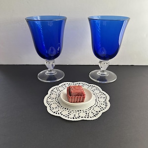 May include: Two cobalt blue stemmed glasses with clear bases and a small white plate with a red square on a white lace doily. The glasses have a flared bowl shape.