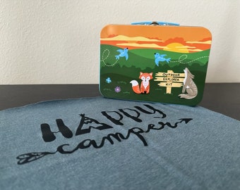 Puede incluir: Una fiambrera de metal azul y naranja con una escena de puesta de sol y el texto "Outdoor Explorer Kit". Un zorro, un lobo y dos pájaros están en la fiambrera. Una camiseta azul con el texto "Happy Camper" está en el fondo.