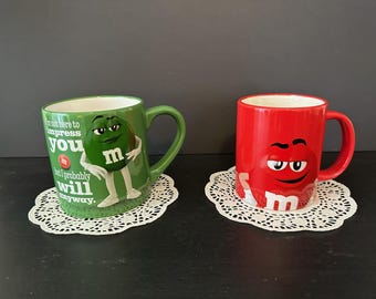 May include: Two ceramic mugs featuring M&M's characters. One is green with the text "I'm not here to impress you but I probably will anyway." The other is red with a smiling M&M's character. Both mugs sit on white doilies.