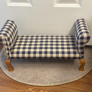 May include: A small, upholstered bench with a navy blue and cream checkered pattern. The bench has curved armrests and wooden legs. It is placed on a round, light gray rug.