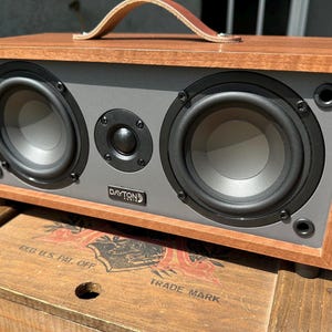 May include: A rectangular wooden speaker with a gray front panel and two large black speakers. It has a brown leather handle and the Dayton Audio logo. The speaker sits on a wooden box.