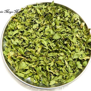 Pode incluir: Uma tigela de metal cheia de folhas verdes secas. As folhas são pequenas e têm uma aparência ligeiramente enrolada. O texto "Organic Mega Mart" é visível no canto superior esquerdo.