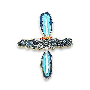 May include: A gold-toned enamel pin in the shape of a cross. The cross is made up of two blue and white shapes that resemble flames, with a black and gray shape in the center. The flames are outlined in gold.