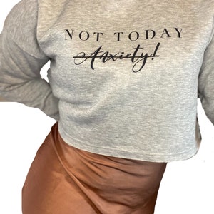 May include: A light gray cropped sweatshirt with the text "NOT TODAY Anxiety!" in black. The sweatshirt has long sleeves and a crew neck. The model is wearing a brown satin skirt.